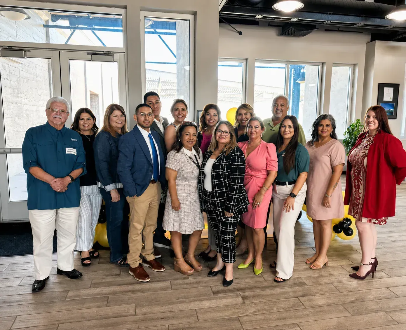 Group of people posing indoors in a bright room with large windows, wearing formal and colorful outfits.