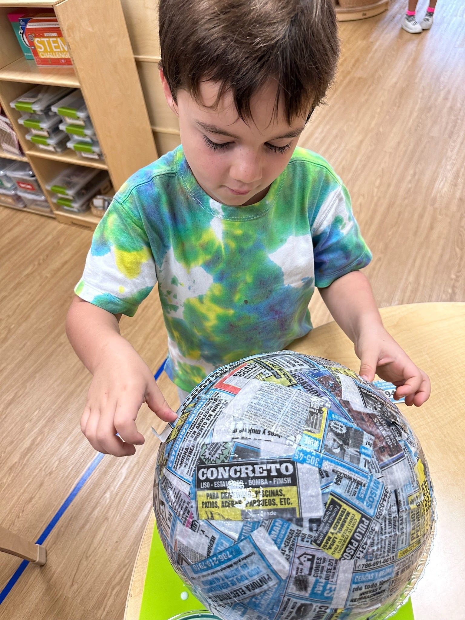 Montessori child in a tie-dye shirt holding a large newspaper-covered ball in a classroom.