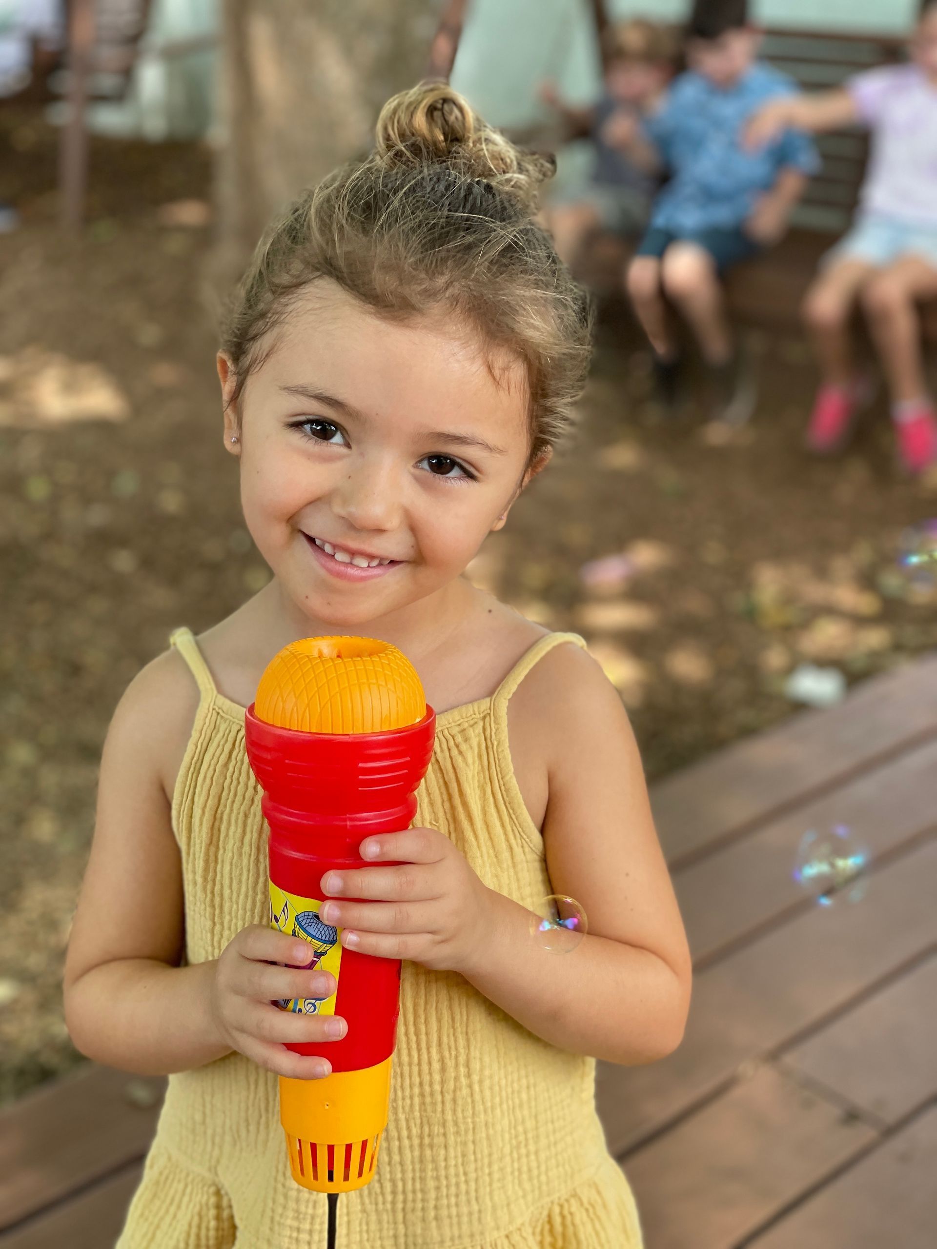 Smiling Montessori child holding a red and yellow toy microphone on a wooden deck outdoors
