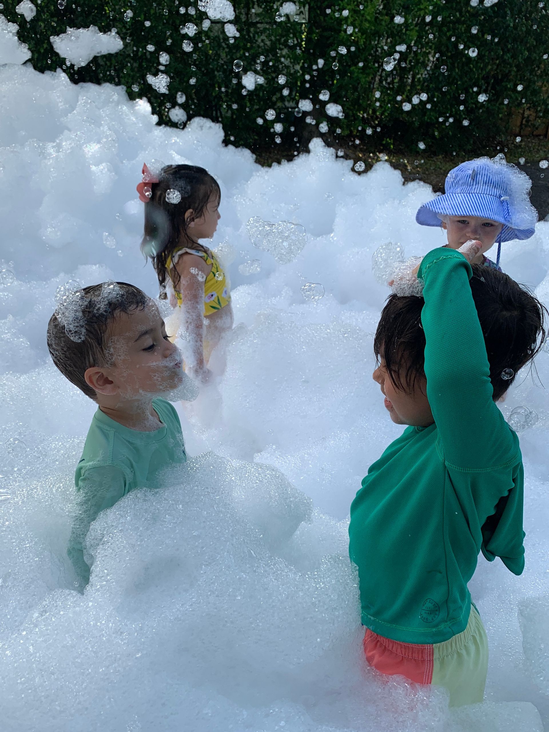 Montessori children playing in thick white foam outdoors, with one child wearing a blue hat.
