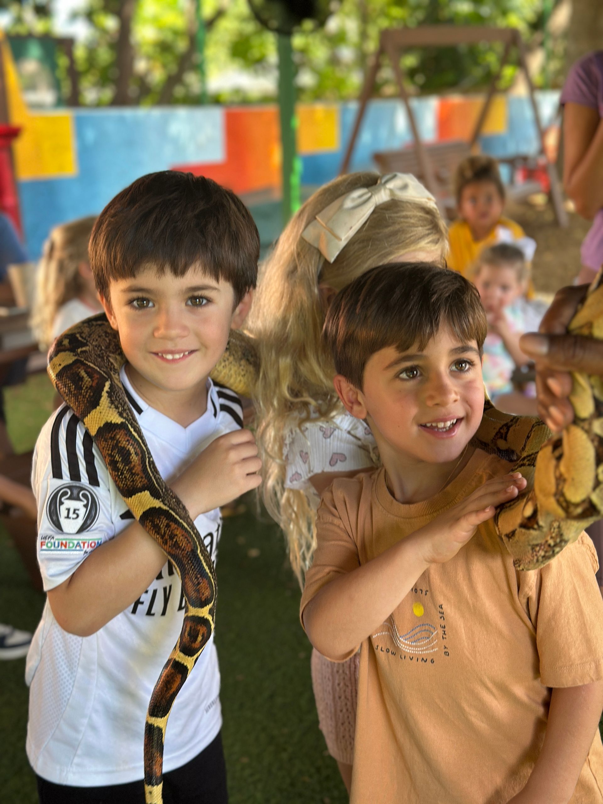 Two smiling Montessori children hold snakes at an outdoor fair, with colorful booths in the background.