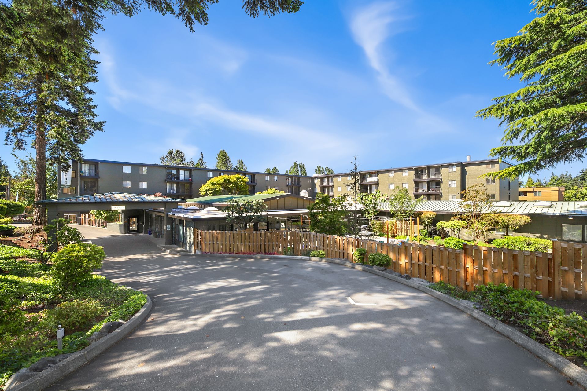 Curved driveway leading to a modern apartment complex under a bright blue sky, framed by trees and wooden fencing