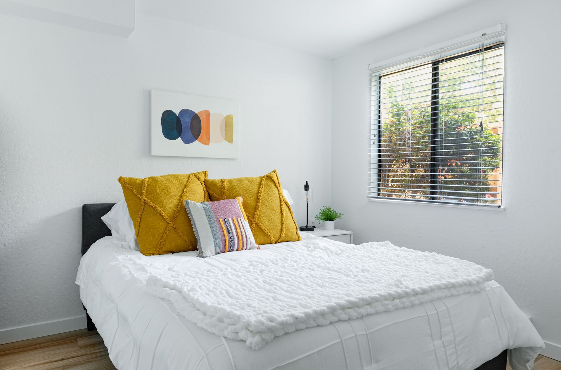 Bright bedroom with a white bed, mustard yellow pillows, wall art, and a window with blinds