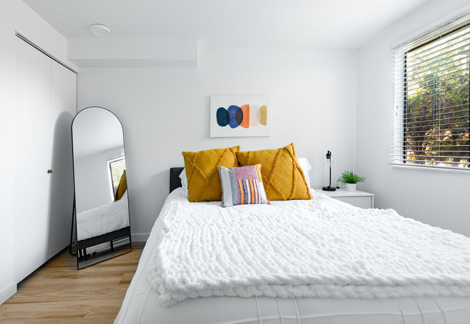 Bright minimalist bedroom with white bedding, yellow pillows, a floor mirror, and a window with blinds.