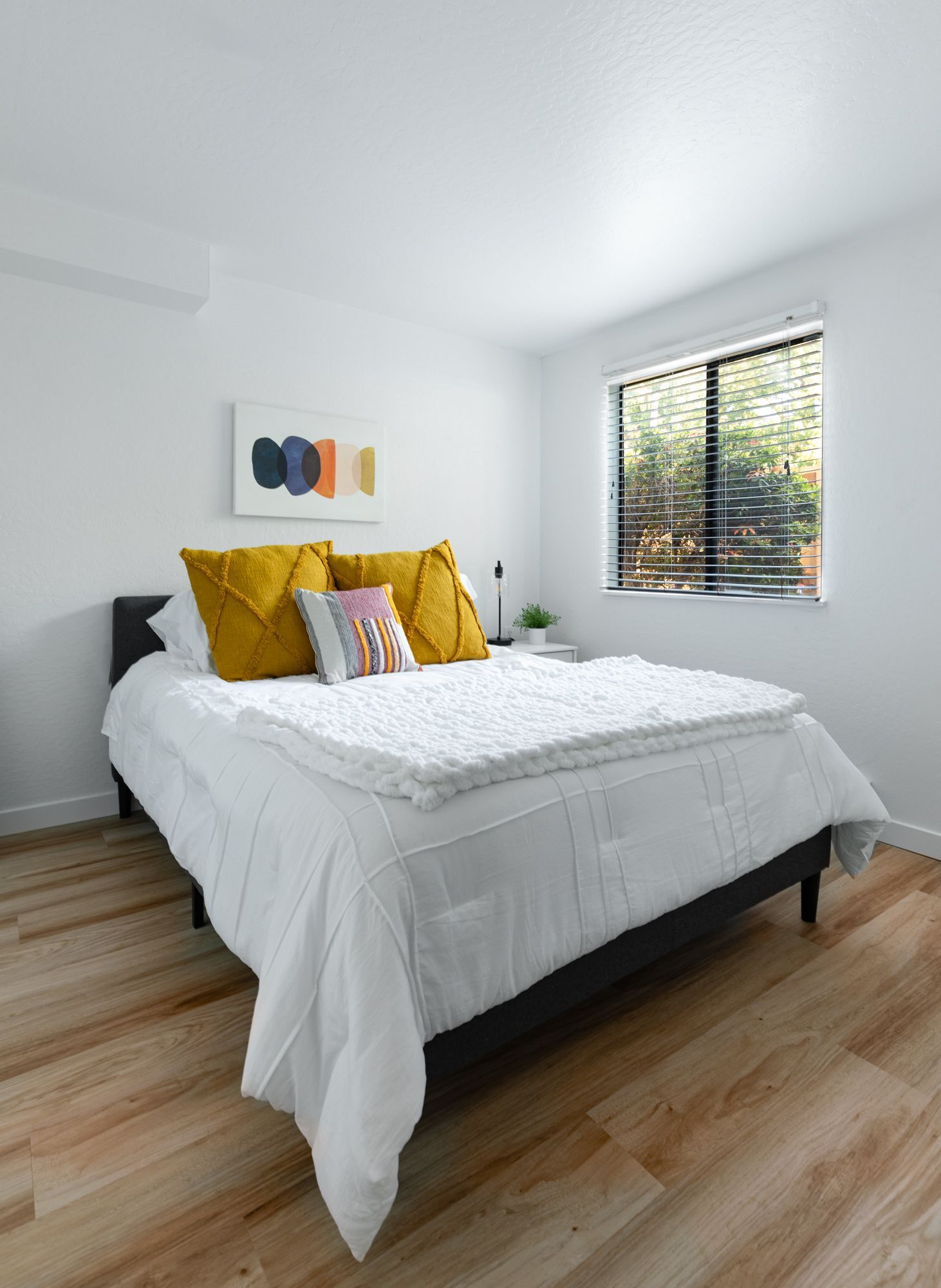 Bright bedroom with a white bed, mustard pillows, wood floors, and a window with blinds