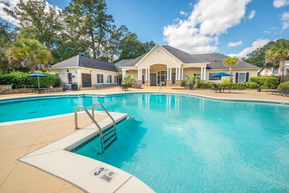 Swimming pool in front of a clubhouse on a sunny day. Blue water, tan deck, and green trees.