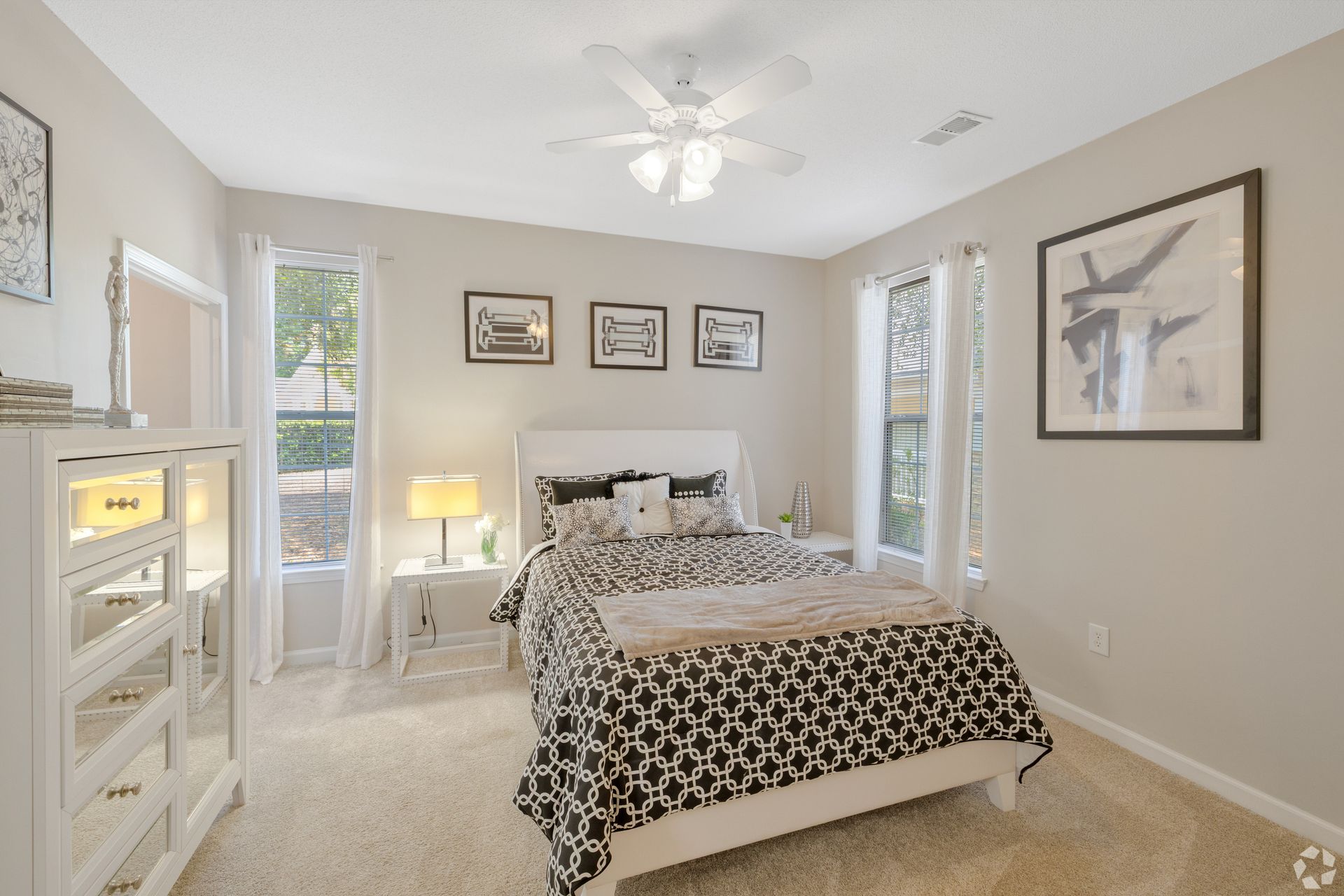 Bright bedroom with a white bed, beige walls, wall art, and a ceiling fan near windows.