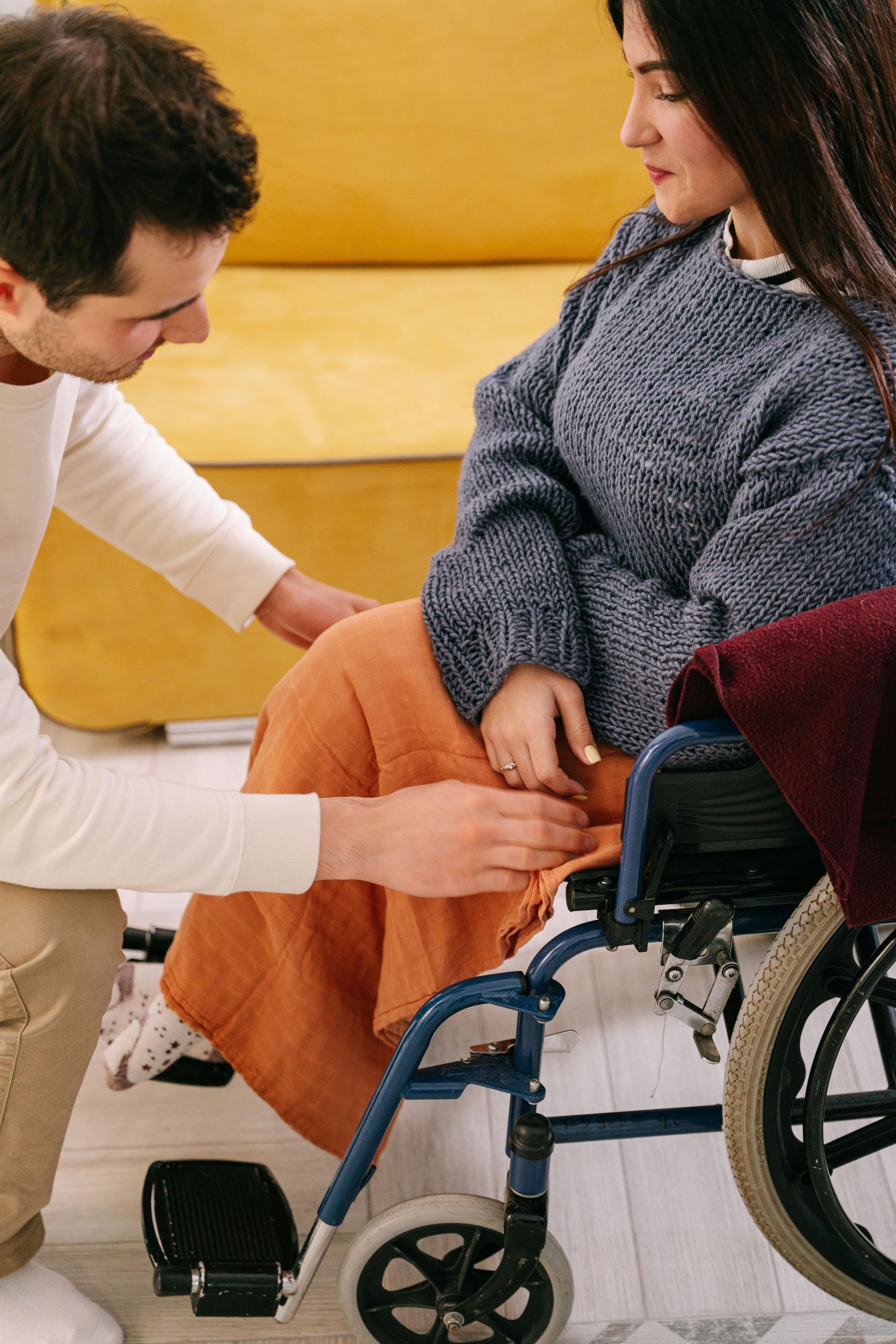 A person kneels to adjust a blanket over the legs of an individual seated in a wheelchair in a brightly lit room.