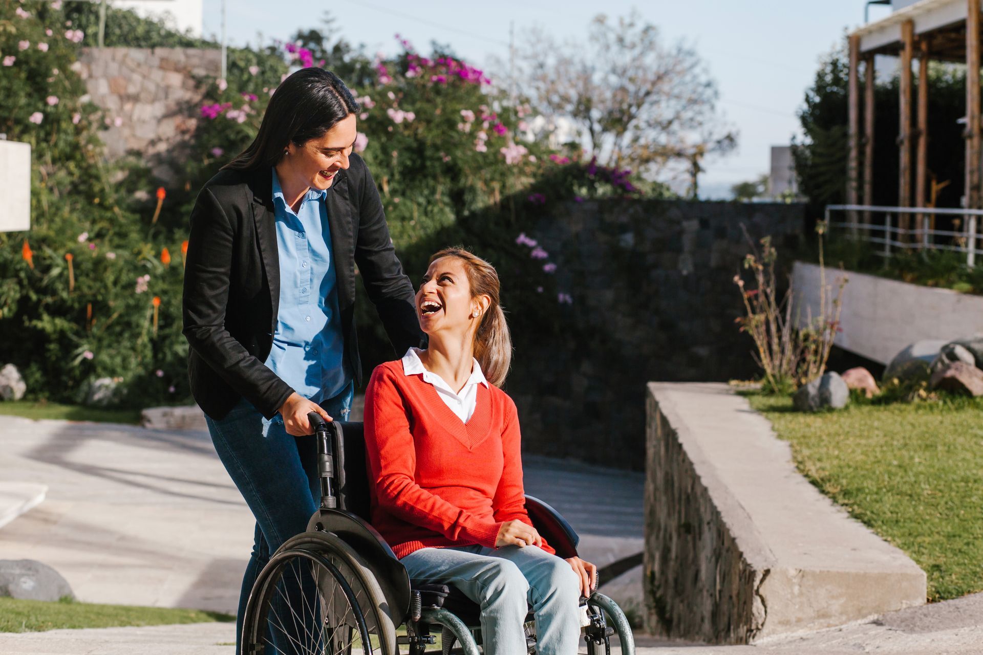 A person in a wheelchair smiles up at a person standing behind them in a sunny, outdoor garden setting.