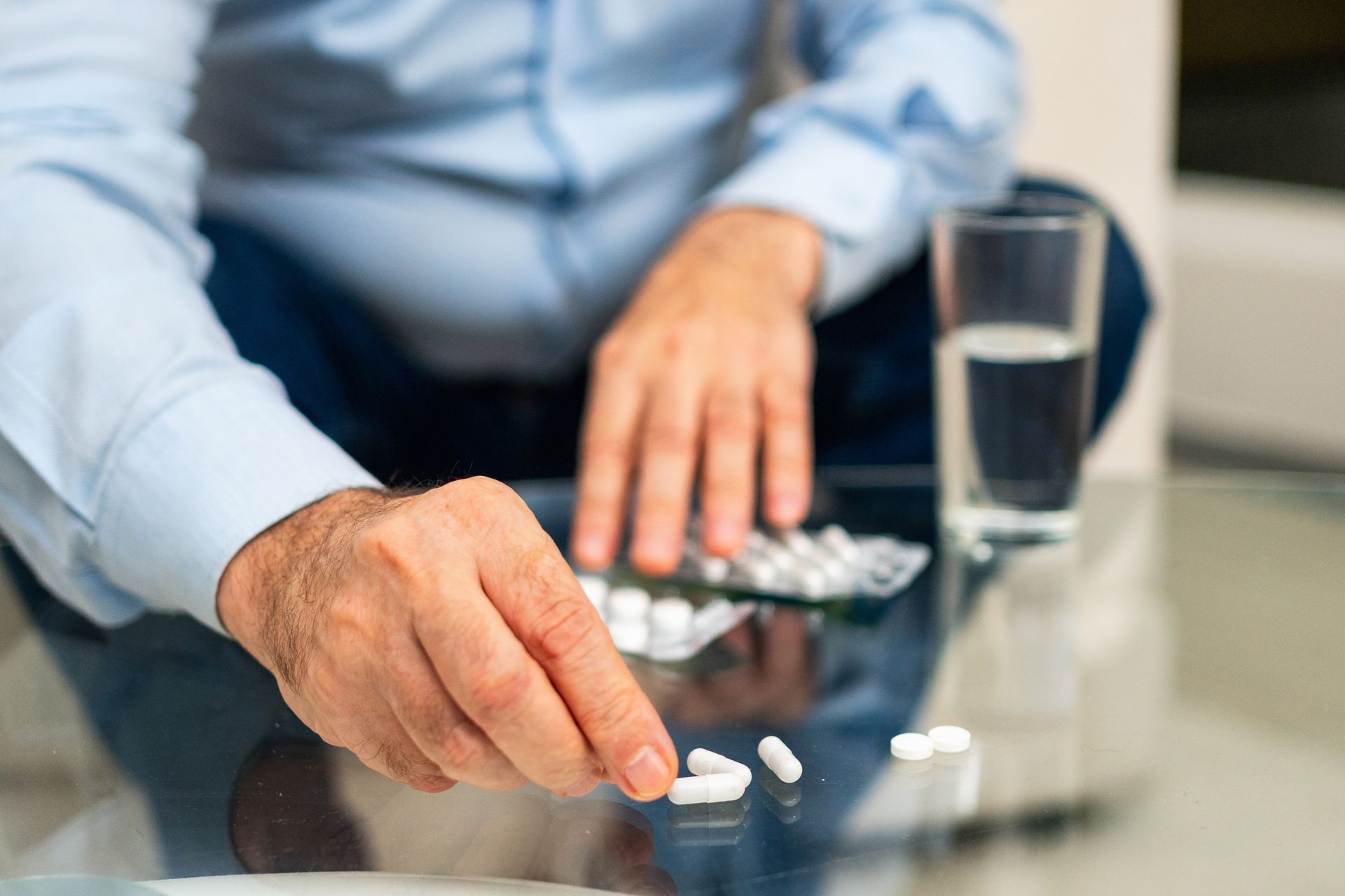 A person in a light blue shirt sitting at a glass table, picking up a white pill next to a glass of water and blister packs.