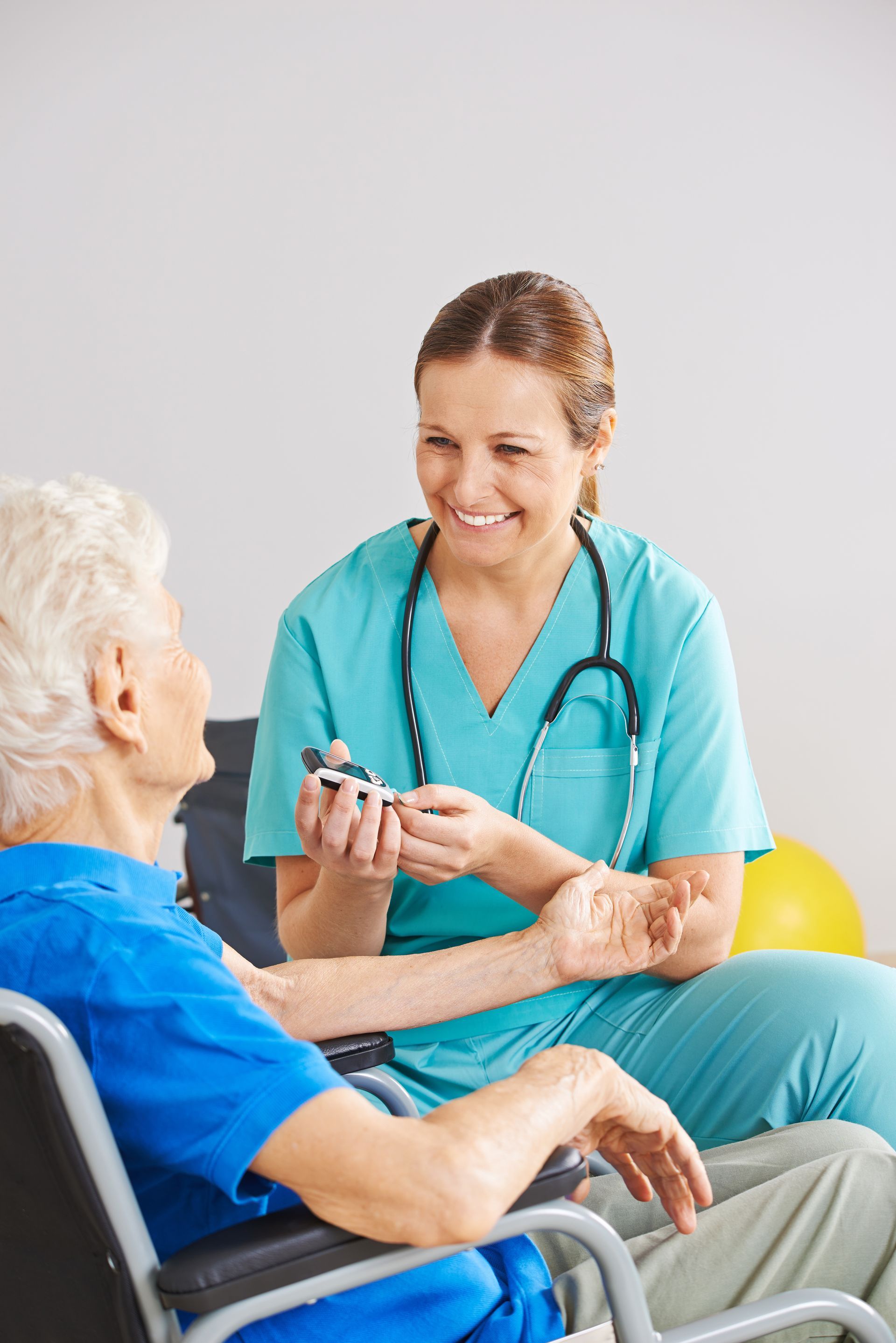 A healthcare professional in teal scrubs smiling while taking an elderly patient’s pulse.