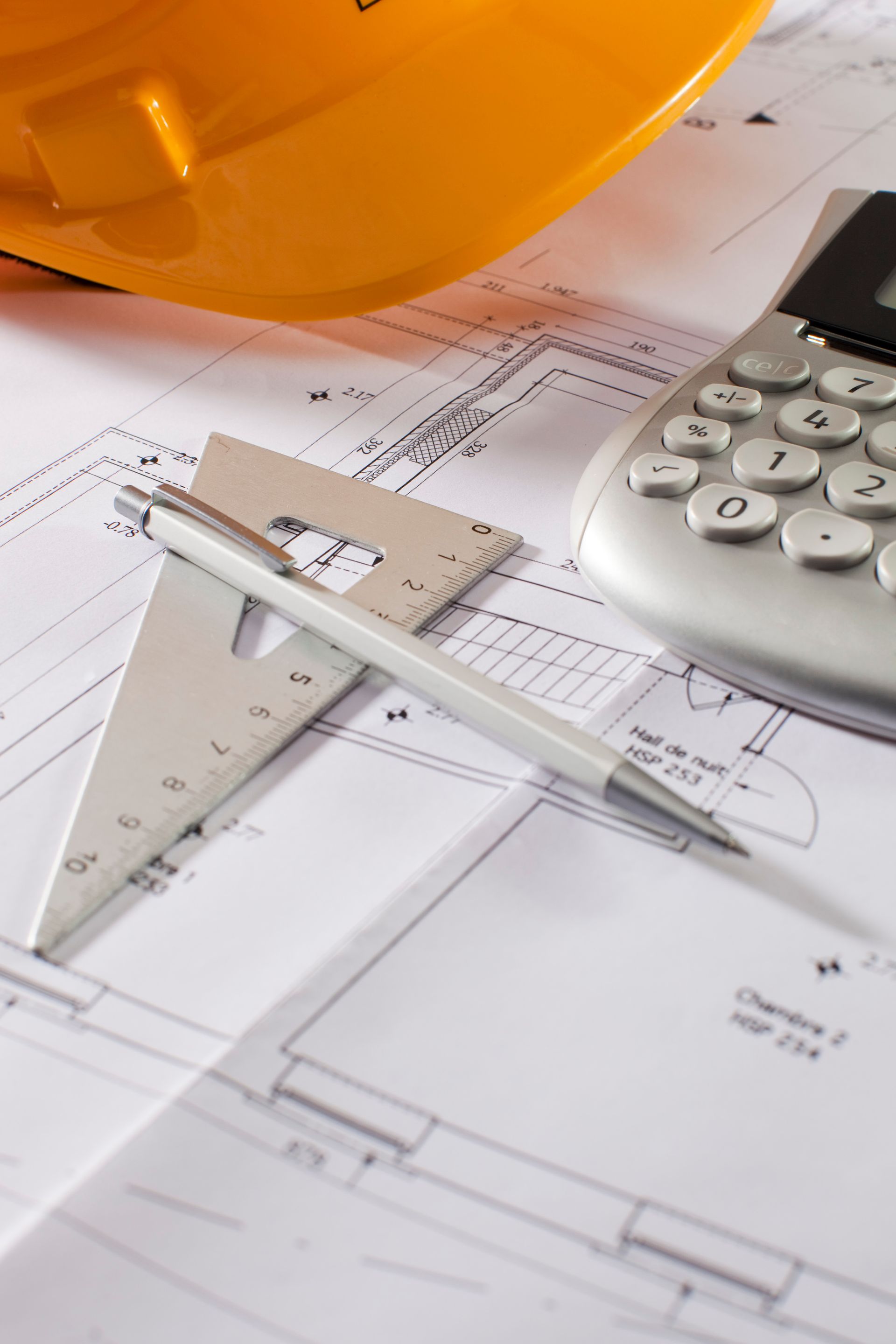 Yellow hard hat, calculator, pencil, and drafting compass on architectural blueprints.
