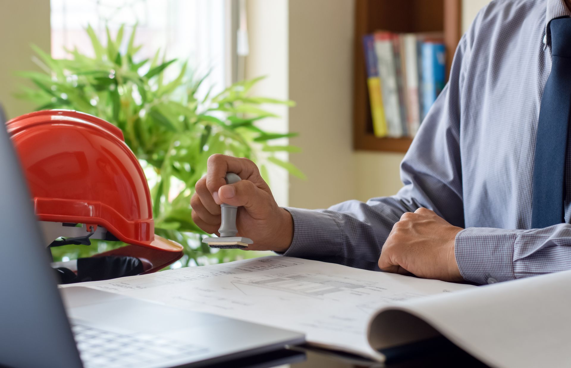 Person stamping paperwork beside a laptop and red hard hat at a desk.