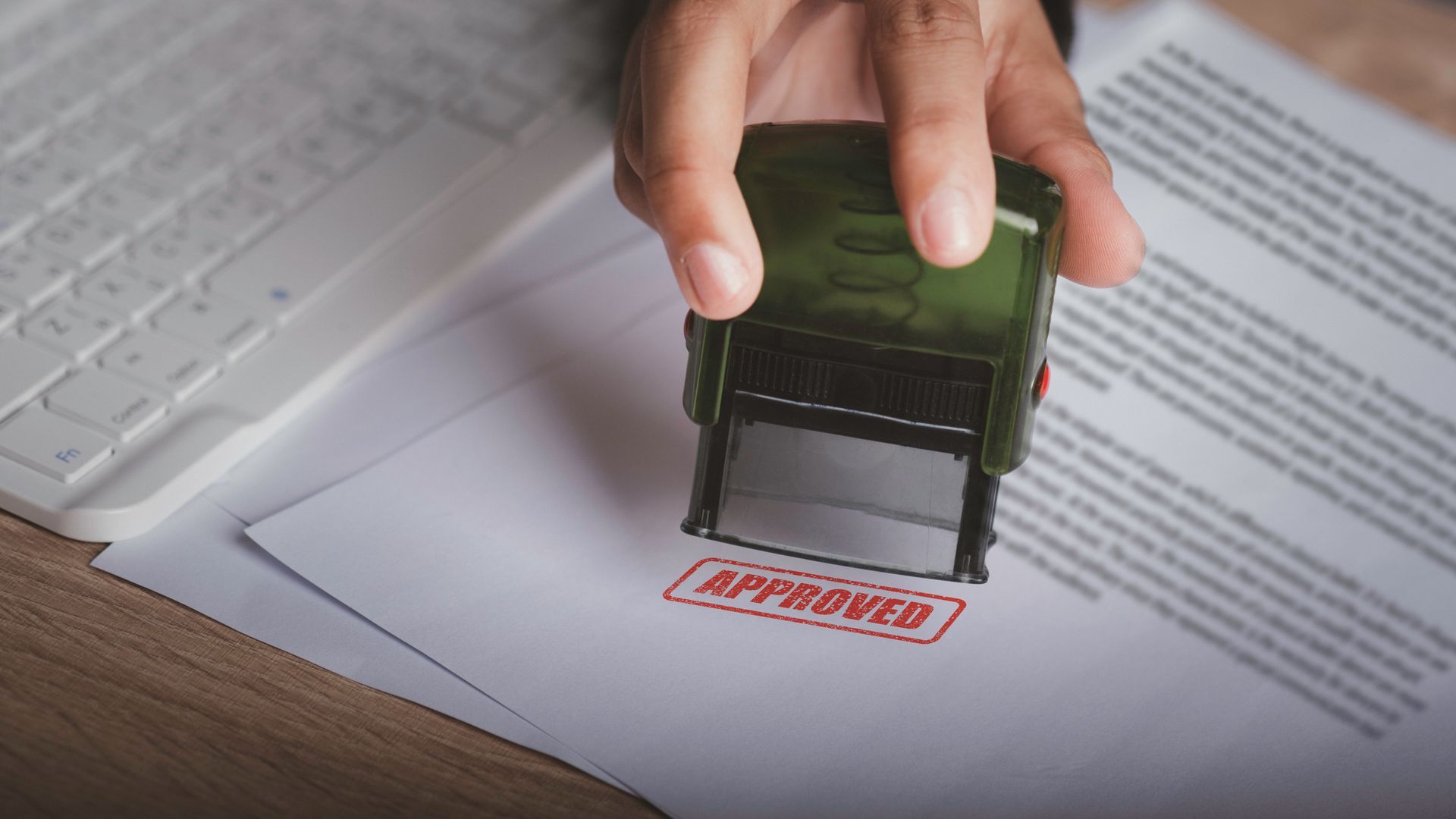 Hand stamping an APPROVED mark on a document on a desk, with a laptop nearby