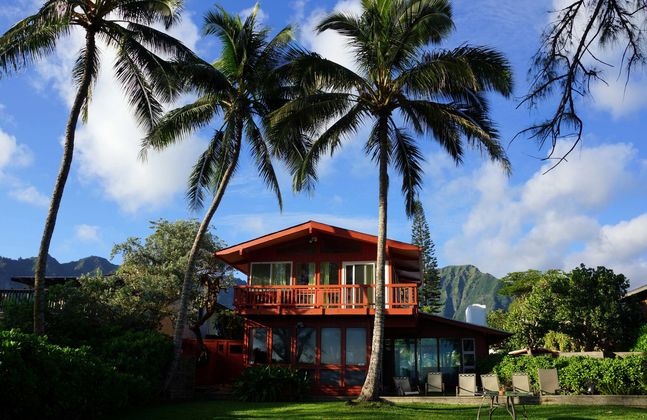 Tropical house with red roof beneath palm trees, set on a lush green lawn with mountains in the background