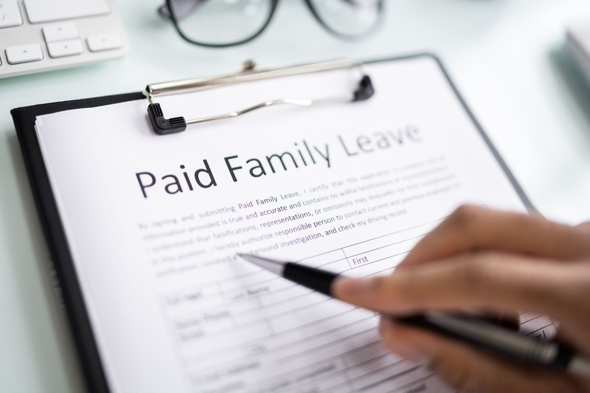Clipboard with “Paid Family Leave” form and a hand holding a pen over the document. Clipboard with “Paid Family Leave” form and a hand holding a pen over the document.