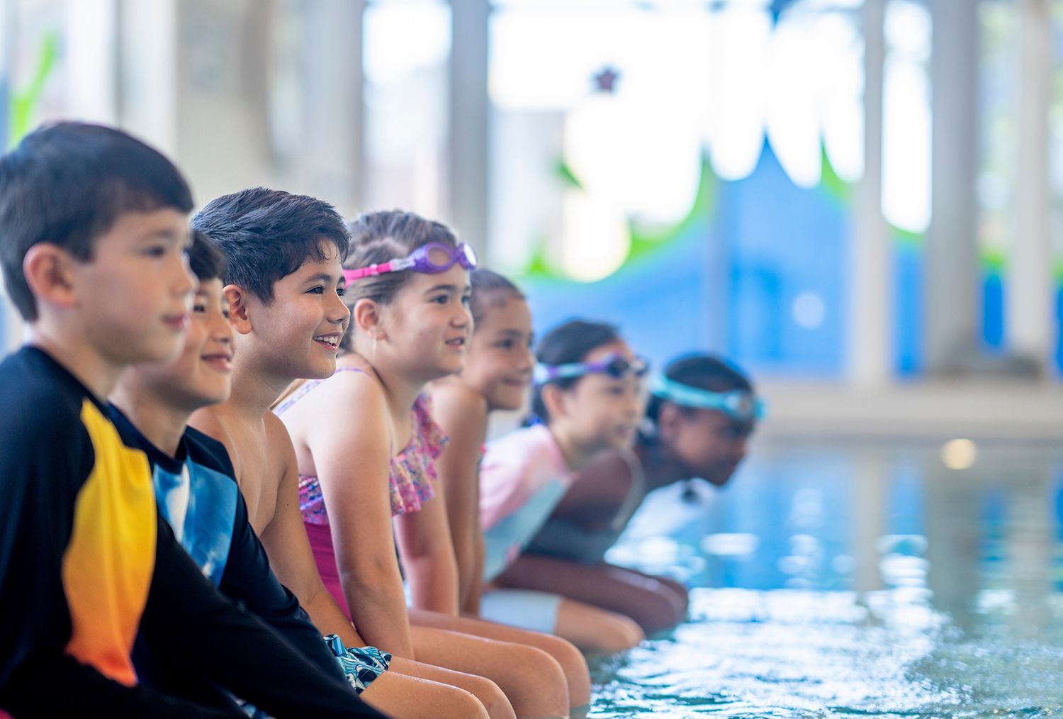 Children sitting at the edge of an indoor pool, smiling and preparing to swim
