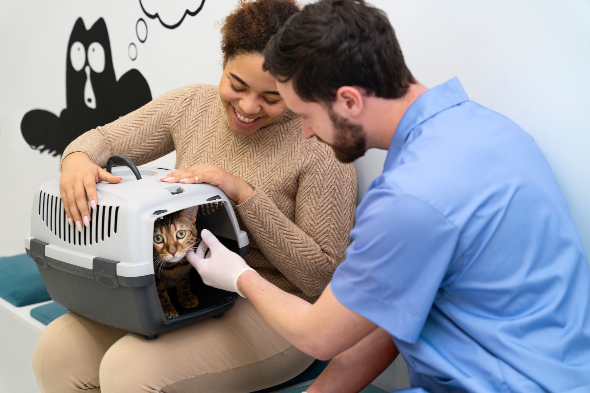 Two people smiling at a cat in a gray pet carrier indoors