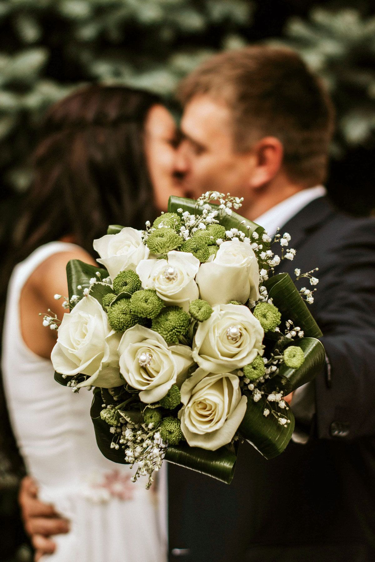 Bride and groom kissing behind a bouquet of white roses and greenery