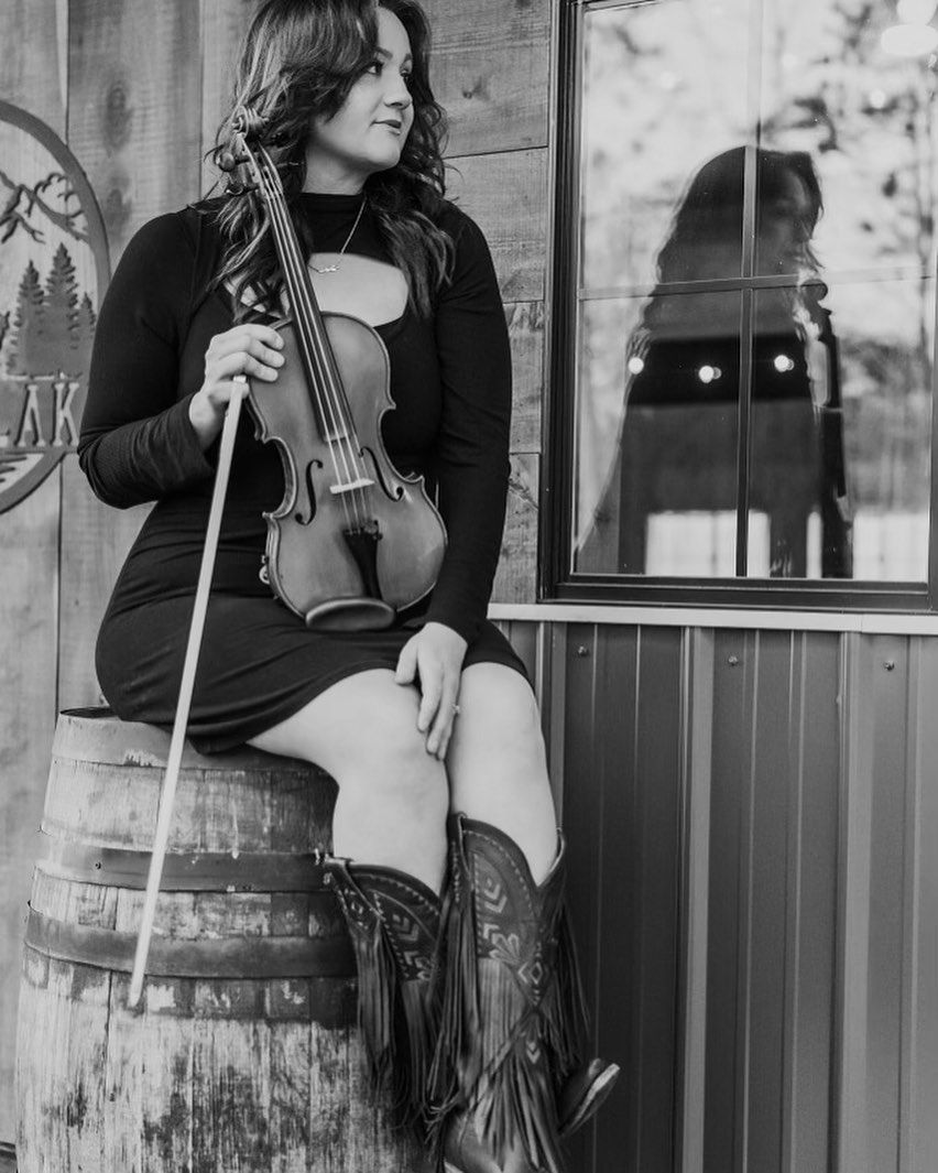 Woman in dark dress holding a violin and bow, seated on a barrel beside a window.