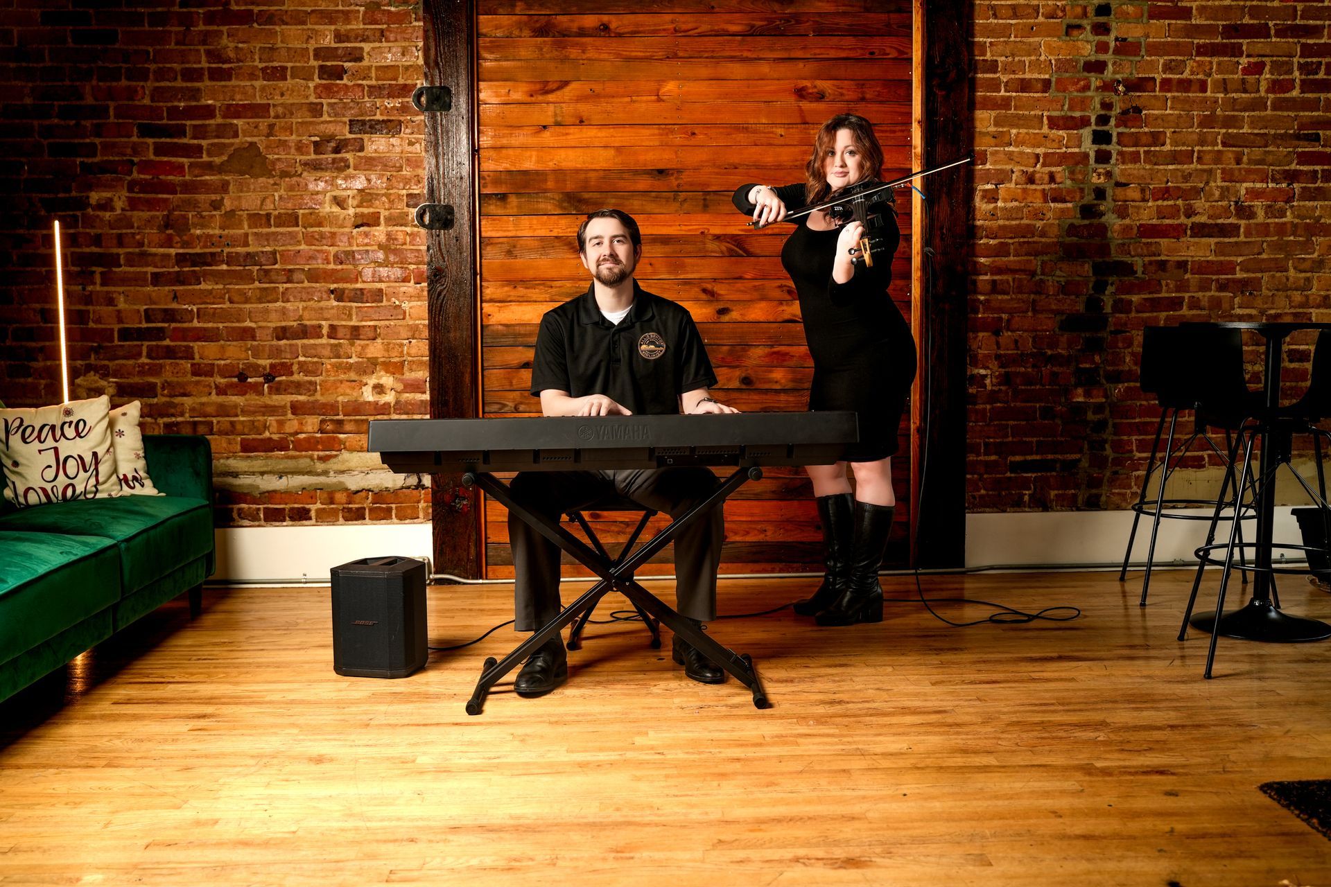 Man seated at a keyboard in a brick-walled studio while a woman stands behind with a camera.