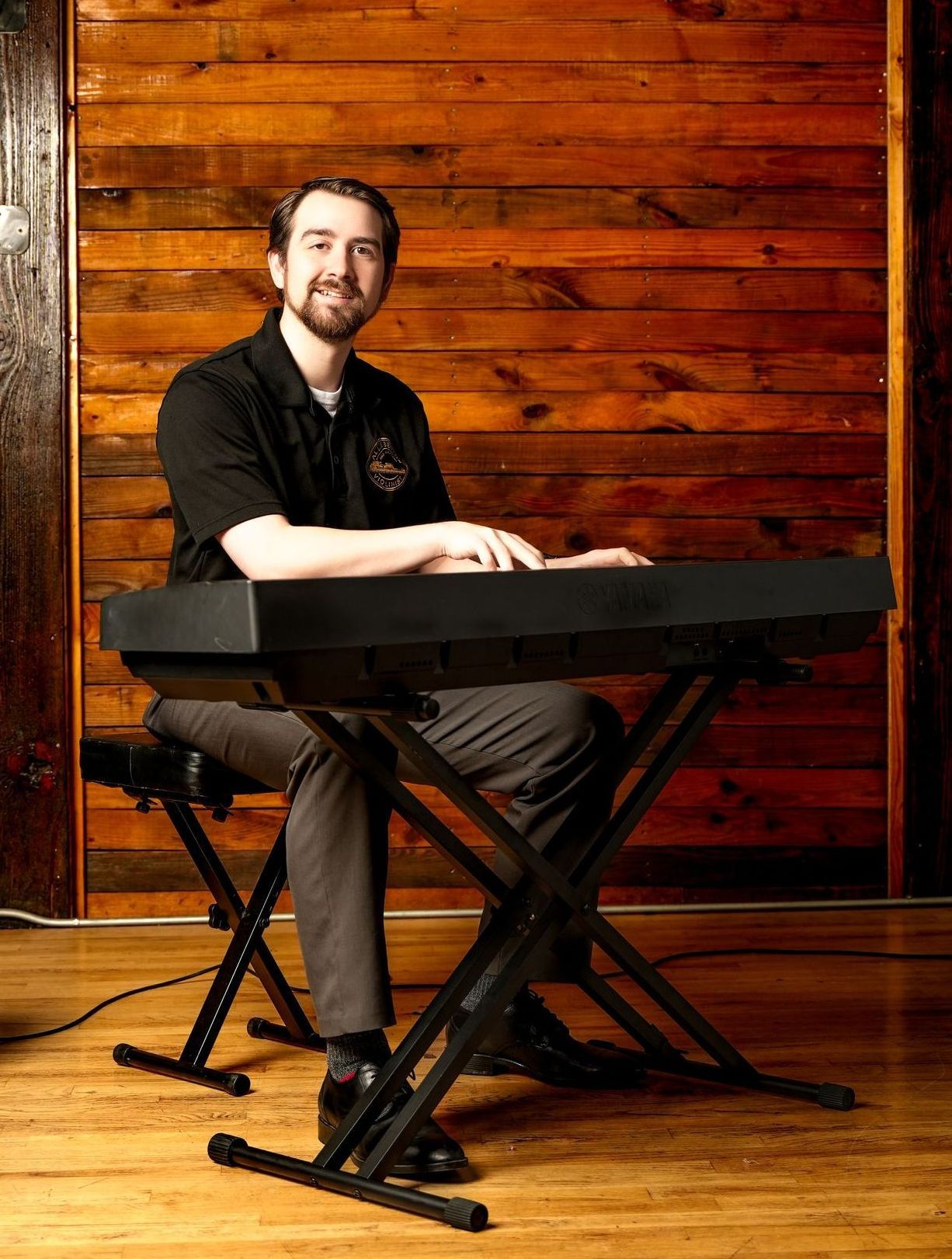 Man seated at an electronic keyboard on a wooden stage, with a warm wood-paneled backdrop.
