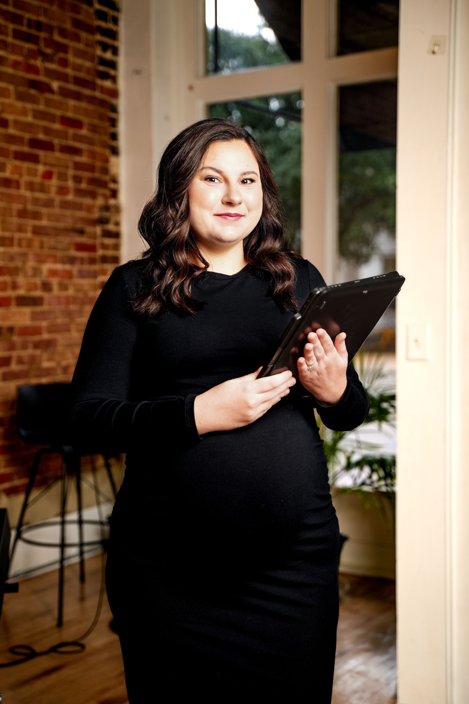 Woman in a black dress holding a clipboard in a sunlit room with brick walls and tall windows