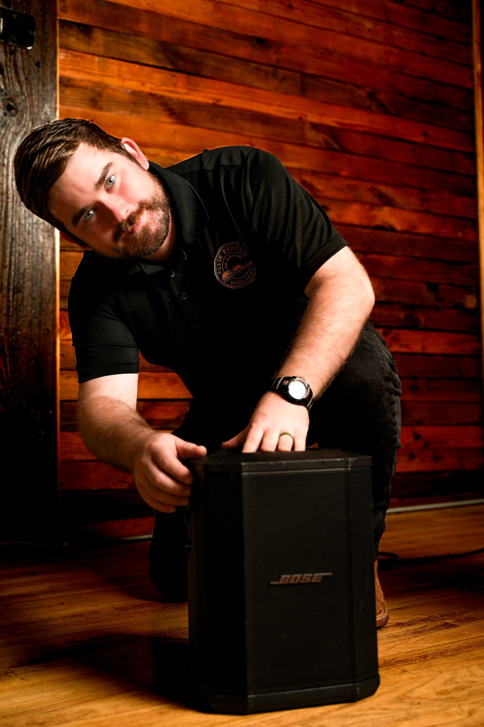 Man crouching beside a black speaker in a wood-paneled room