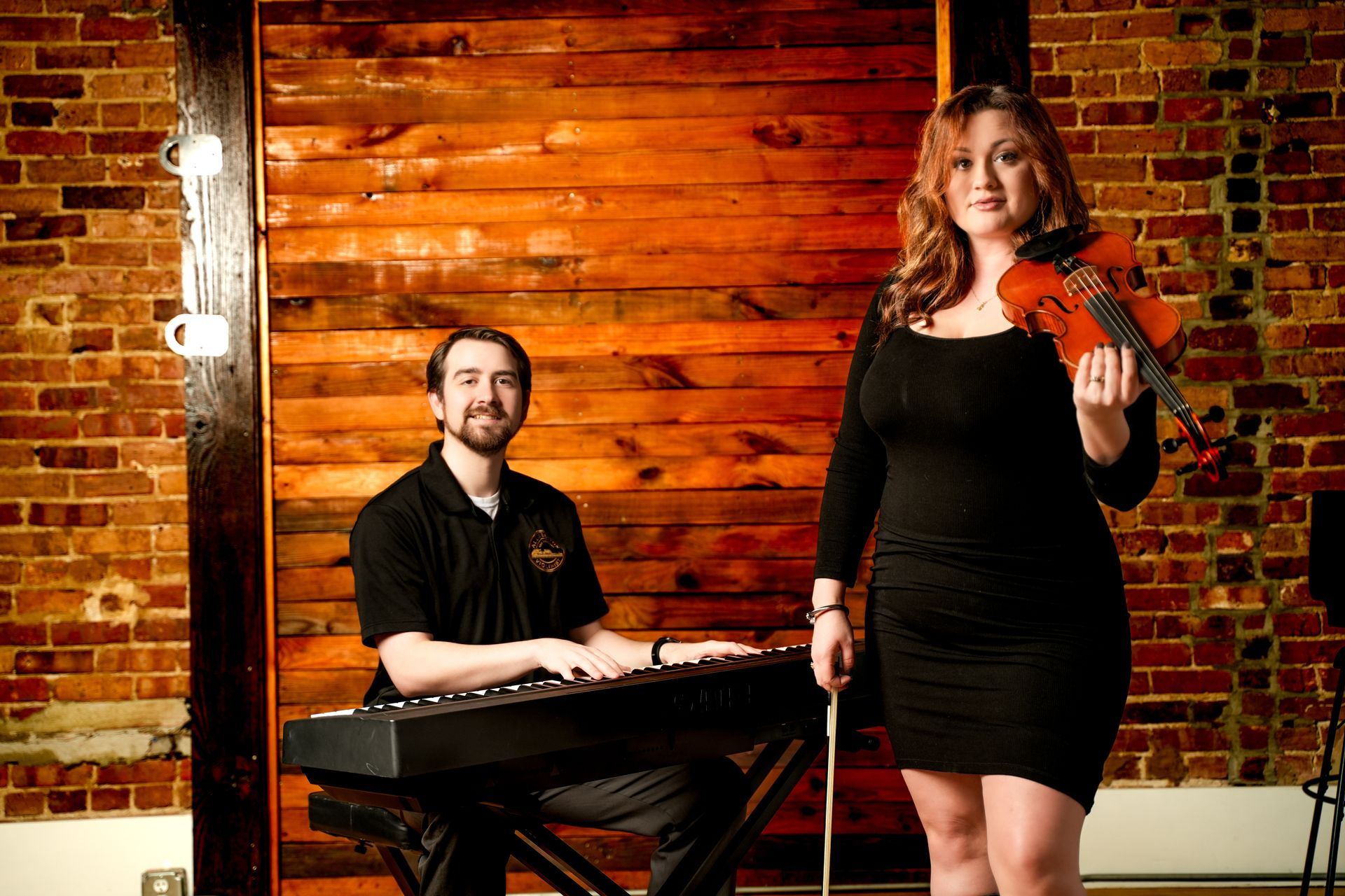 Musicians performing indoors: pianist at keyboard and violinist in black dress before a wooden wall.
