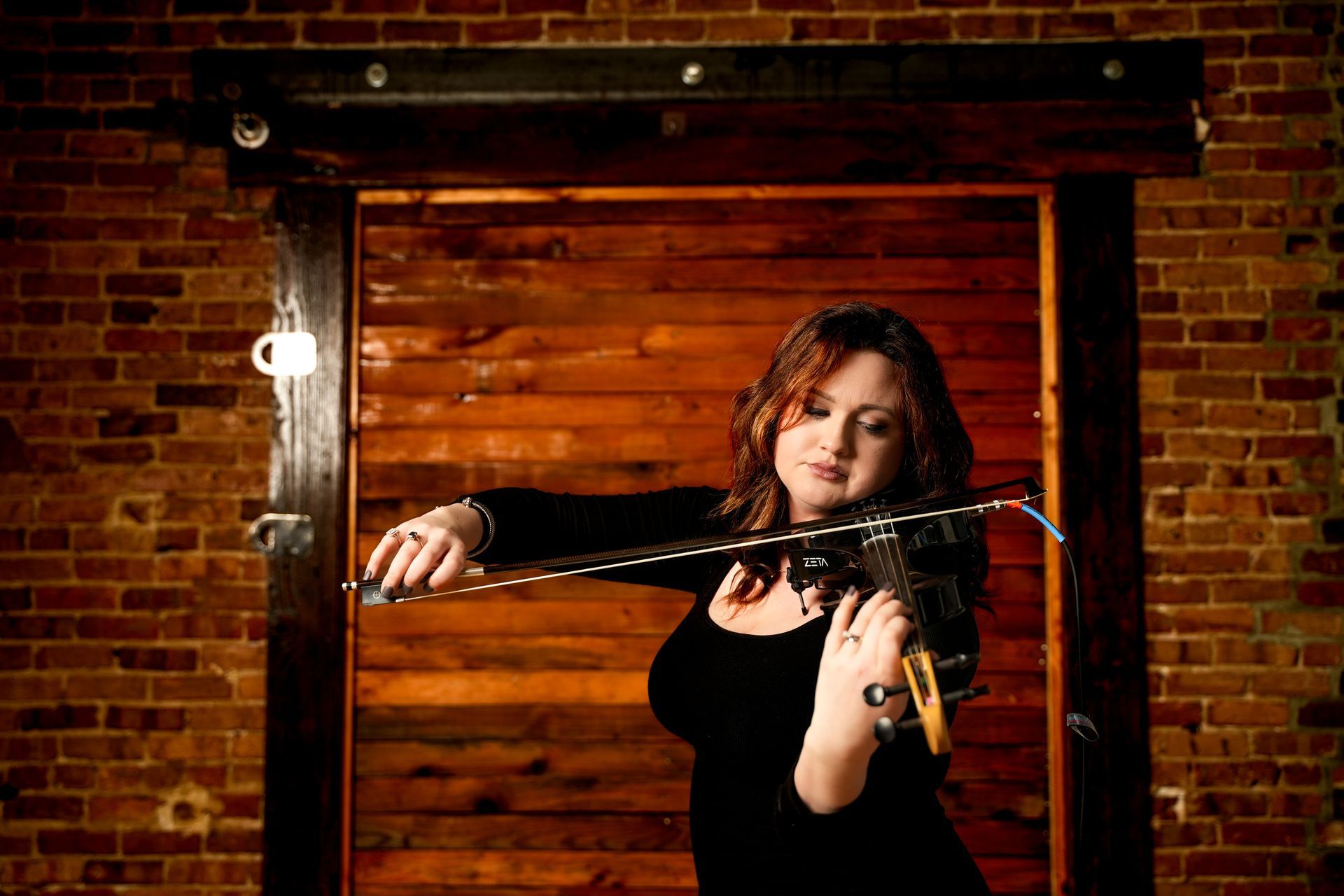 Woman in black dress playing violin in front of a wooden door and brick wall