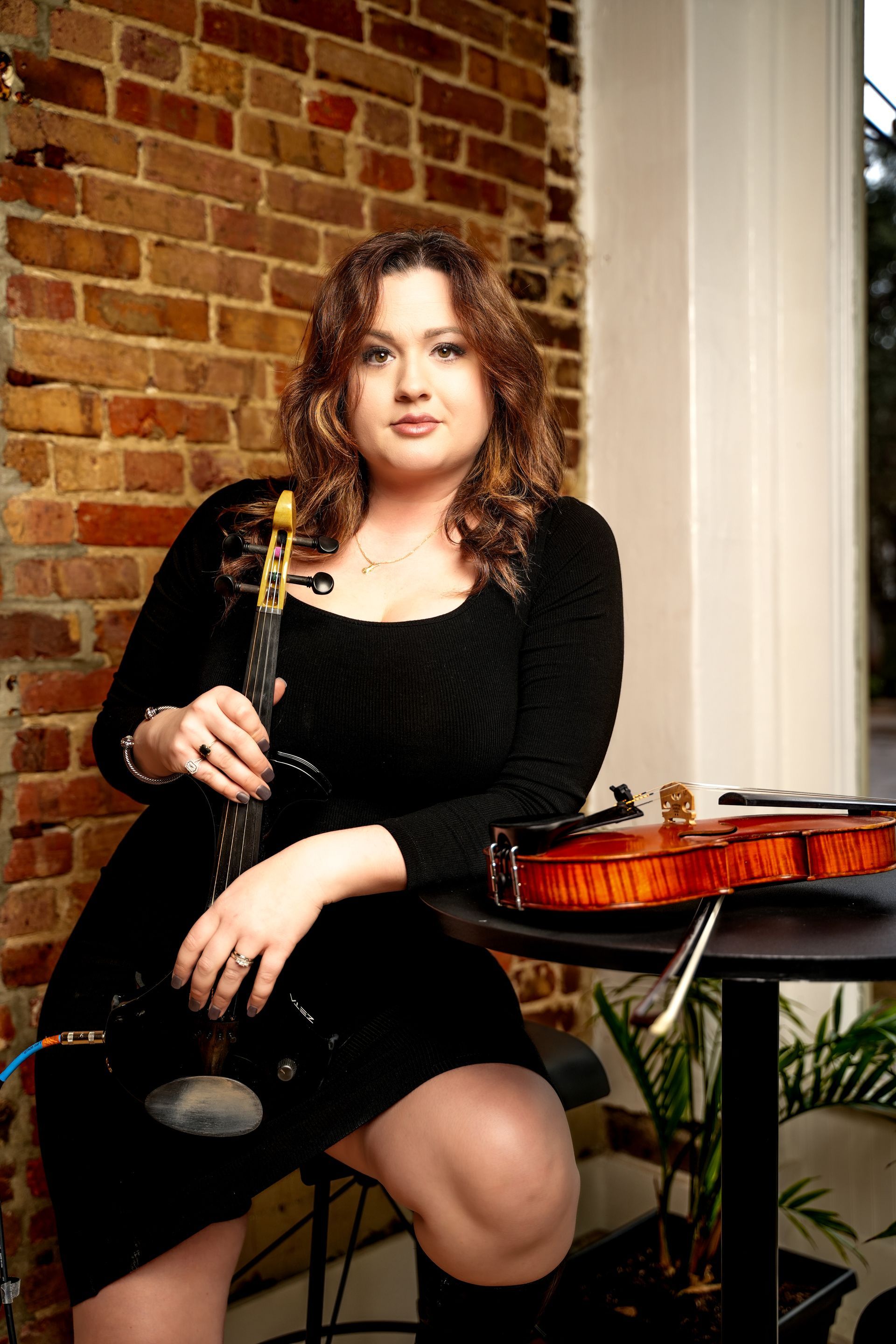 Woman seated with a violin and bow beside a brick wall and window, wearing a black dress.