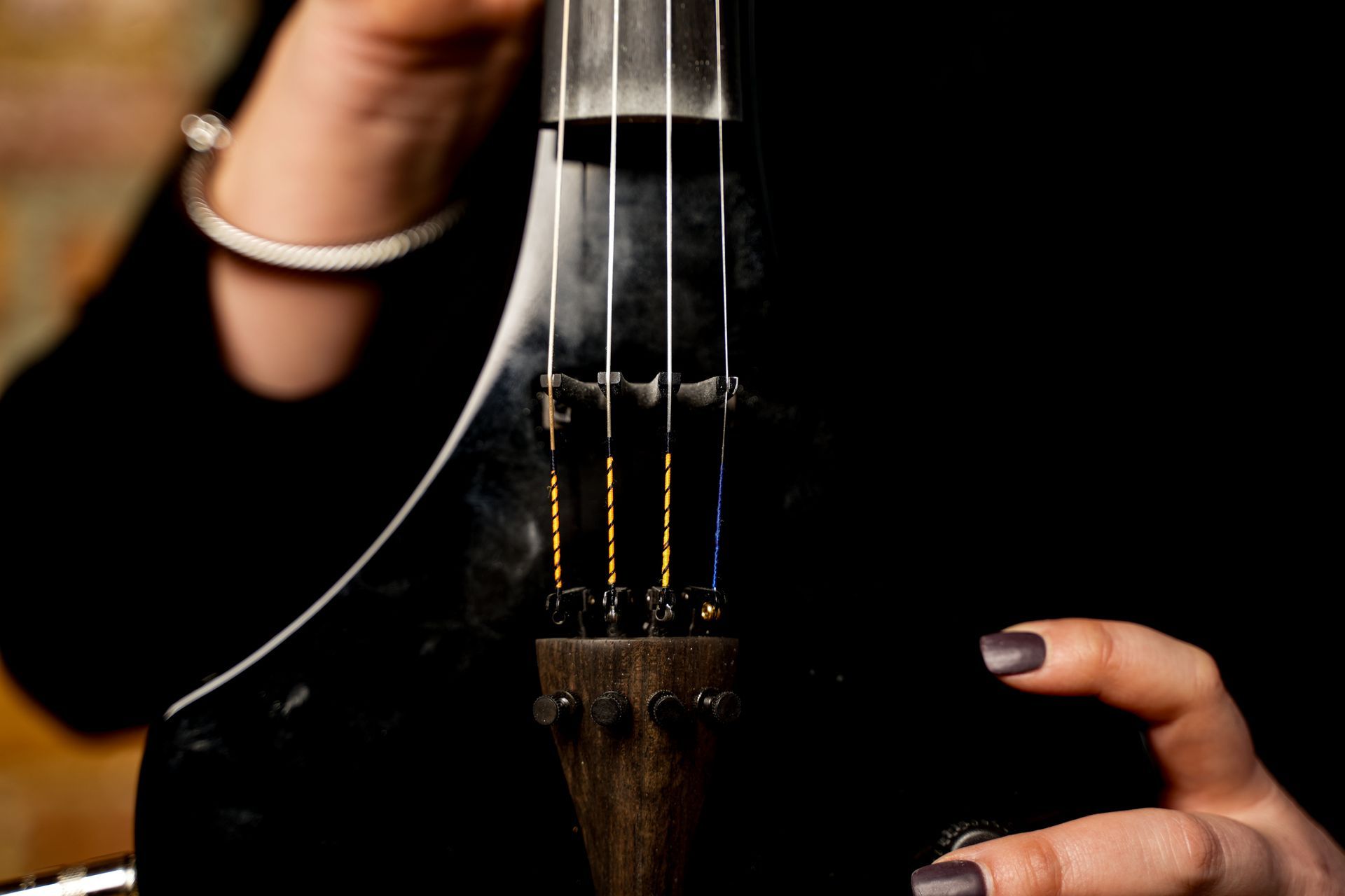 Hands pluck a black string instrument against a dark background, with a bracelet visible on one wrist.