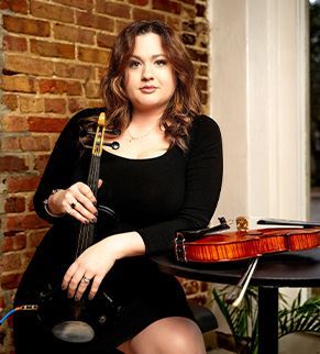 Woman seated with a violin and bow beside a table holding another violin, against a brick wall.