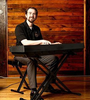 Man seated at a keyboard in front of a wooden wall, smiling in a warm studio setting