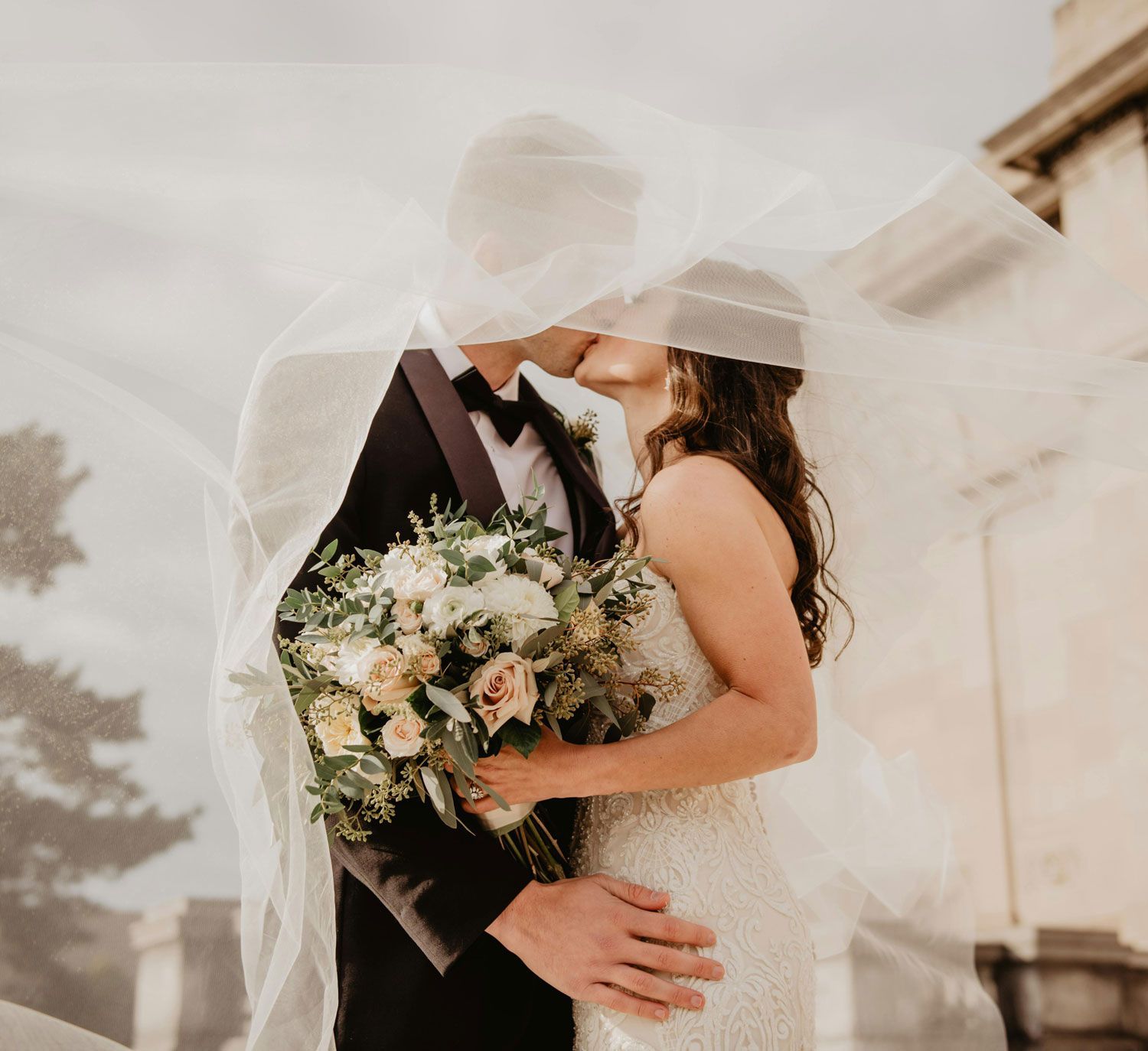 Bride and groom kissing under a flowing veil, holding a bouquet at an outdoor wedding ceremony