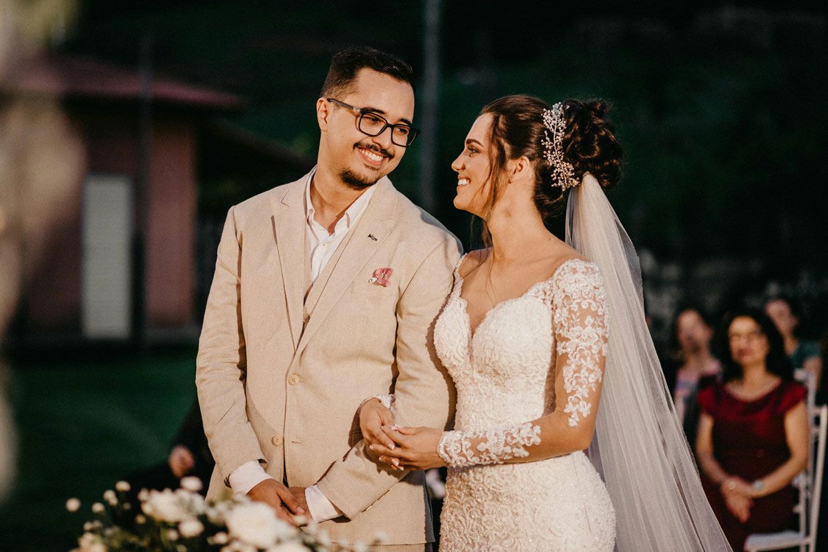 Smiling couple holding hands at a wedding, with the bride in a white lace gown and veil, groom in a beige suit.