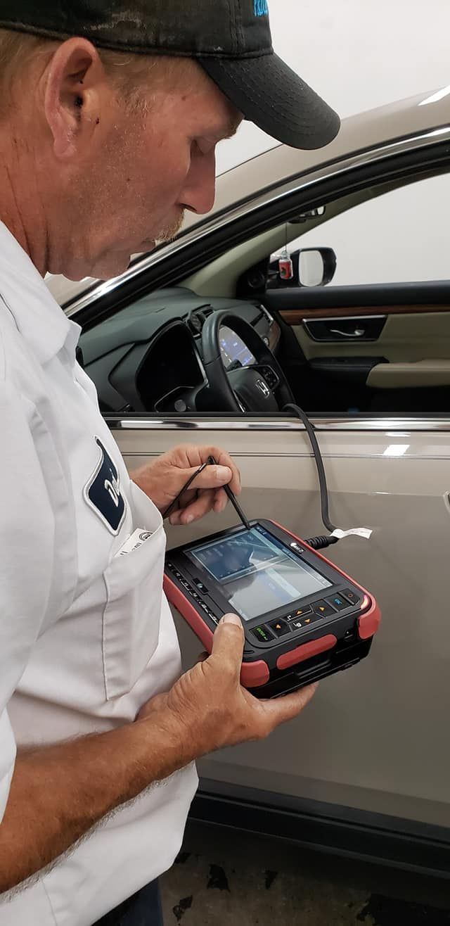 Mechanic using a diagnostic tool on a car. He's wearing a cap and white shirt, looking at the screen.