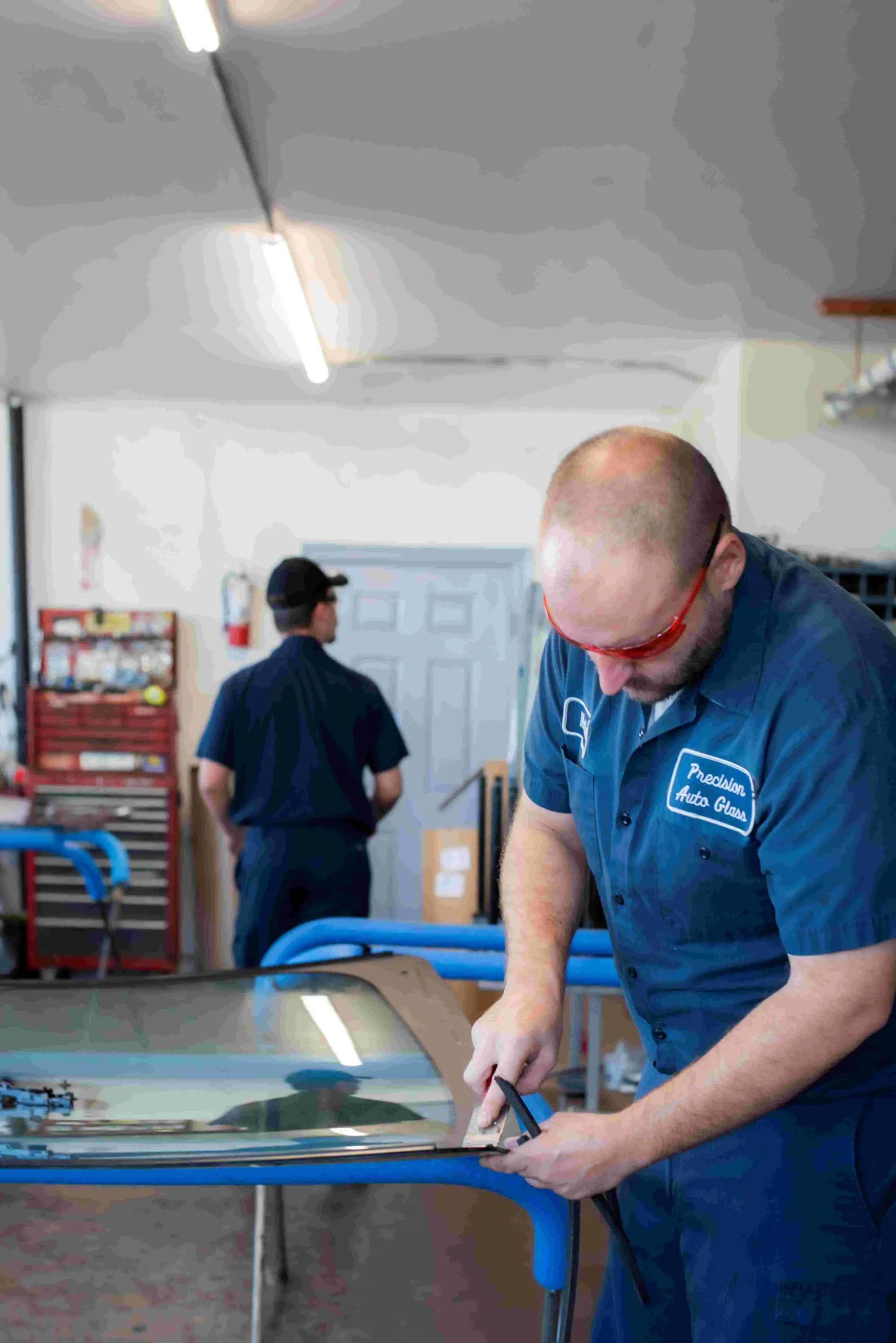 Mechanic in blue uniform cuts metal at a repair shop; another worker in the background.