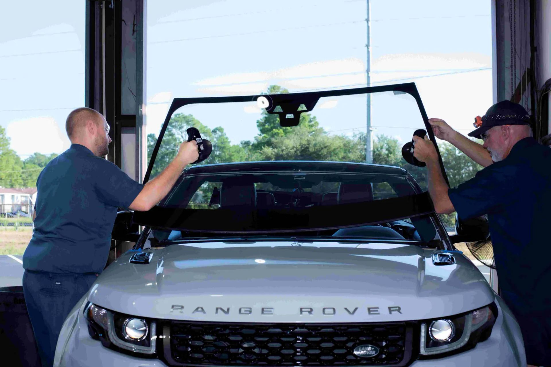 Two men replacing the windshield of a white Range Rover SUV inside a garage.