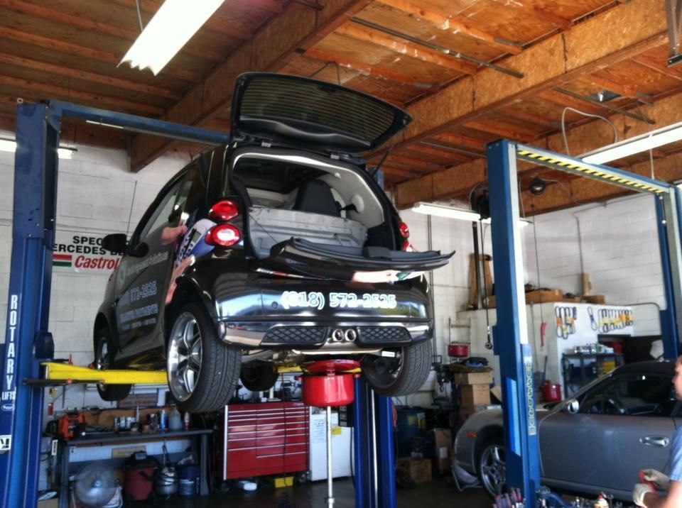 Black Smart car on a lift in a garage with the trunk open.