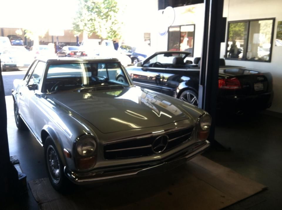 Silver Mercedes convertible in a garage, other cars visible, sunny day.