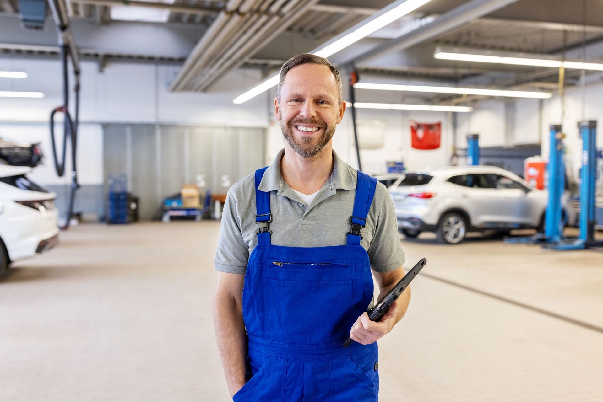 Smiling mechanic in blue overalls, holding tablet, in a car repair shop with vehicles.