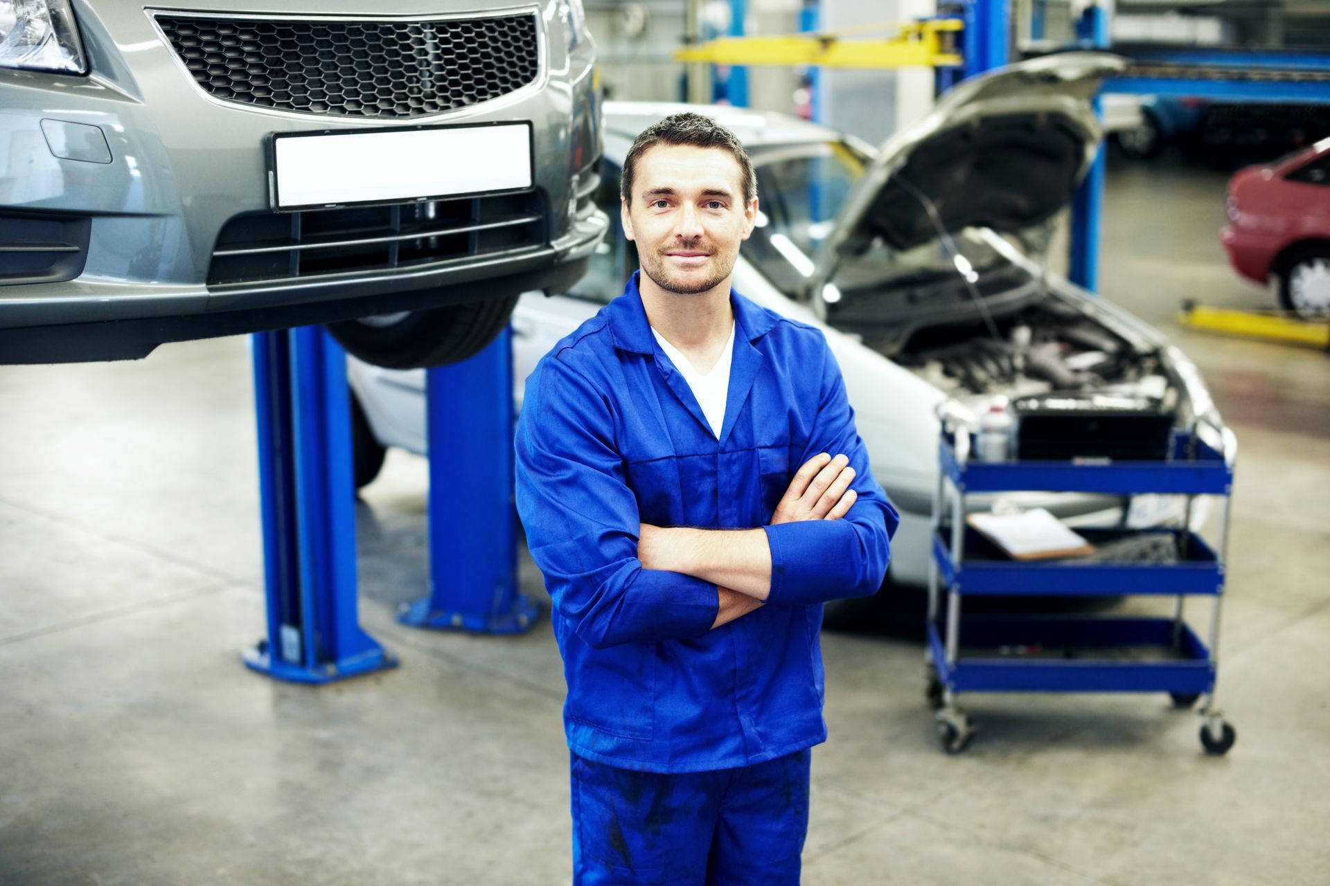 Mechanic in blue uniform smiles, arms crossed, in auto repair shop; car on lift in background.