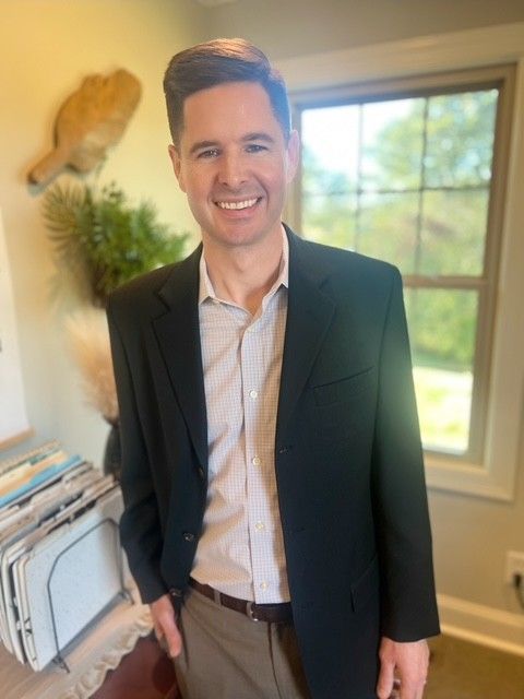 Smiling person in a dark blazer stands indoors by a window in a bright room