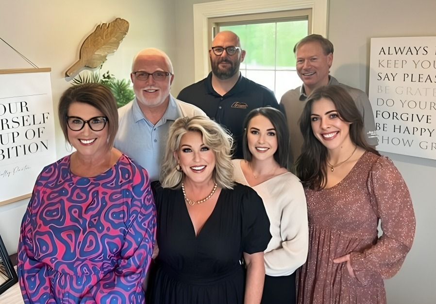 Six smiling adults posing indoors in front of framed wall art.