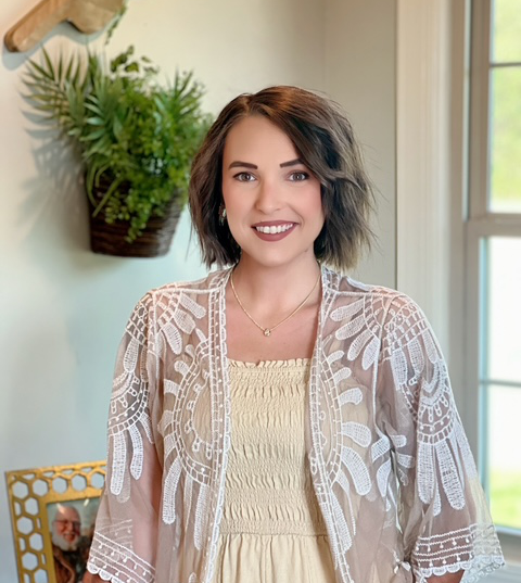 Smiling woman in a cream lace top standing indoors beside a window and hanging plant