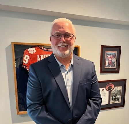 Smiling man in a navy suit standing in an office with sports memorabilia on the wall