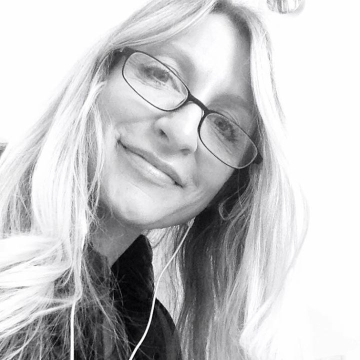 Woman with long blonde hair, glasses, and earphones smiling slightly.
