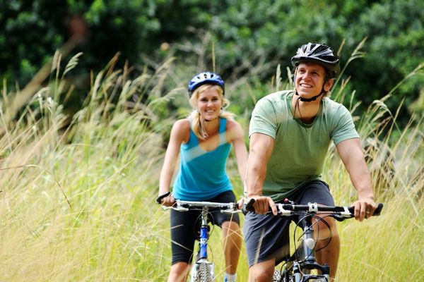 A smiling couple bikes on a path through tall grass, both wearing helmets; the woman in a blue top, the man in a green one.