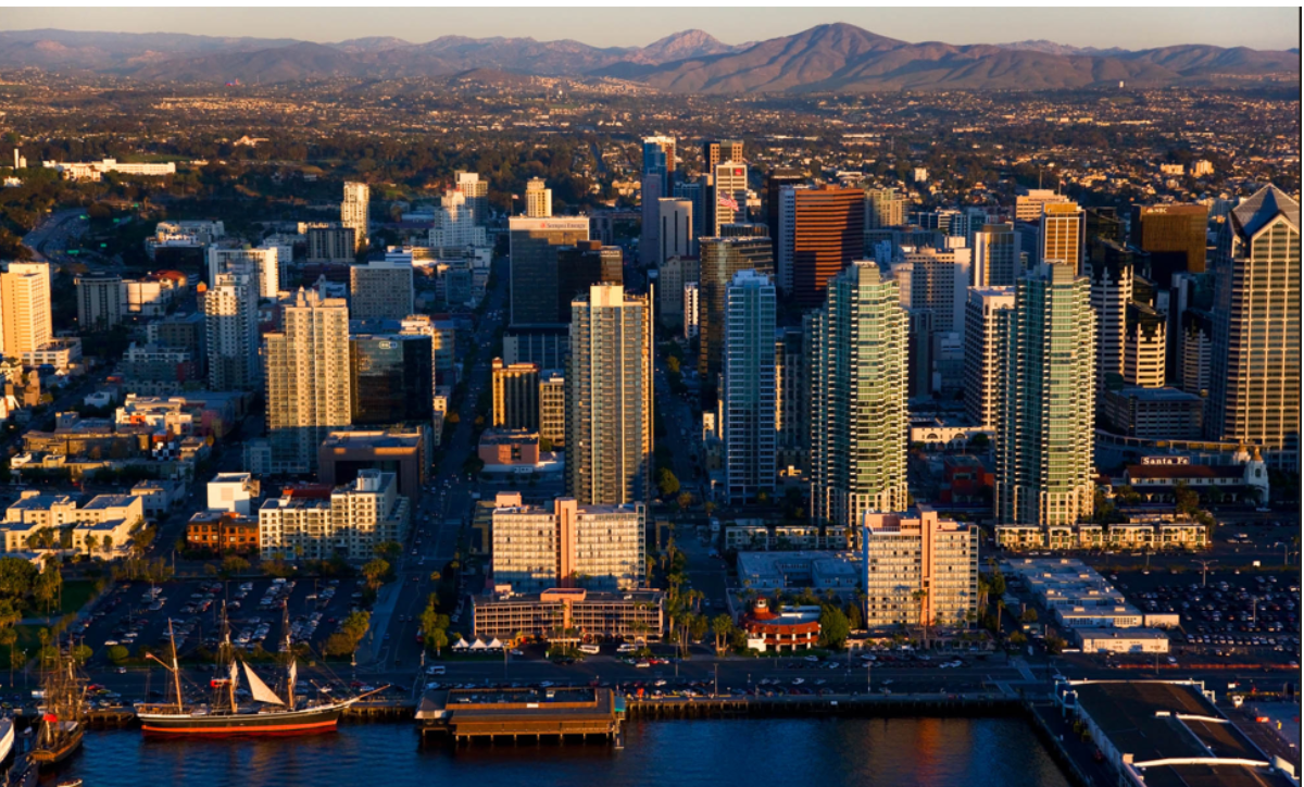 Aerial view of a coastal city skyline with tall glass buildings at sunset, mountains in the distance.